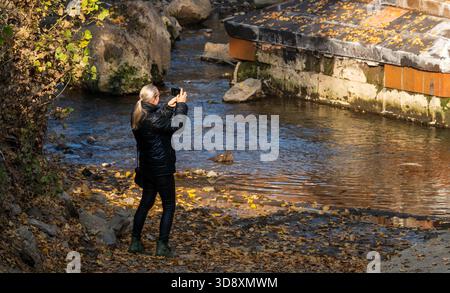 Jerewan Armenien 10.25.2025. Eine Frau fotografiert einen Fluss. Das Wasser ist ruhig und die Blätter auf dem Boden sind braun Stockfoto