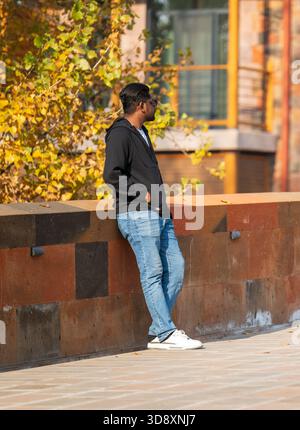 Jerewan Armenien 10.25.2025. Ein Mann in schwarzer Jacke und blauer Jeans lehnt sich an eine Ziegelwand. Er trägt eine Sonnenbrille und sieht aus Stockfoto