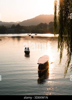 Blick auf Boote, die ruhig auf dem glitzernden Wasser schwimmen, das den goldenen Sonnenuntergang reflektiert, mit fernen Bergen und Trauerweide, Hangzhou, Zhejiang, C Stockfoto