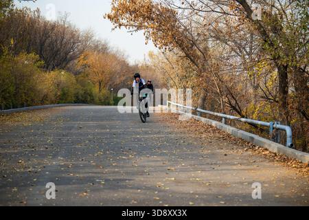 Jerewan Armenien 11.25.2025. Ein Mann fährt mit dem Fahrrad eine Straße hinunter mit Bäumen im Hintergrund. Die Blätter auf den Bäumen ändern ihre Farbe, sodass die s Stockfoto