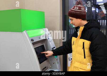Junger Mann in leuchtend gelber Winterjacke und Hut, der einen ATM-Touchscreen bedient, Geld abhebt oder einzahlt. Hochwertige Fotos Stockfoto