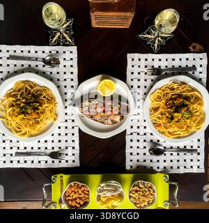 Eine Tischansicht von oben zeigt eine Mahlzeit für zwei Personen, Spaghetti mit Meeresfrüchten und einen gebackenen Fisch in der Mitte. Die Gerichte werden mit Gläsern Wein serviert. Stockfoto