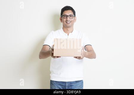 Servicekonzept für Kurierzustellung. Indischer Mann schickt eine braune Box, steht auf einem schlichten Hintergrund mit Schatten. Asiatisches, gutaussehendes Männermodell Stockfoto