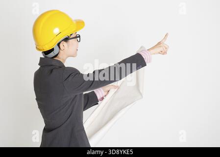 Porträt einer asiatischen Ingenieurin mit einem Schutzhelm, der blauen Druck hält und wegzeigt, auf einem einfachen Hintergrund steht Stockfoto