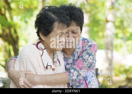 Freimütiger Schuss einer reiferen asiatischen Frau umarmt und tröstet ihre schreiende alte Mutter im Outdoor-Naturpark Stockfoto