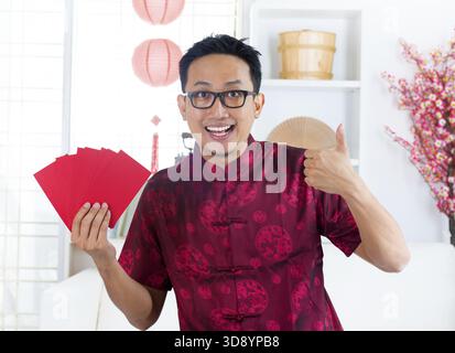 Daumen hoch asiatischer Chinese mit vielen roten Päckchen/ang Pow, drinnen Stockfoto