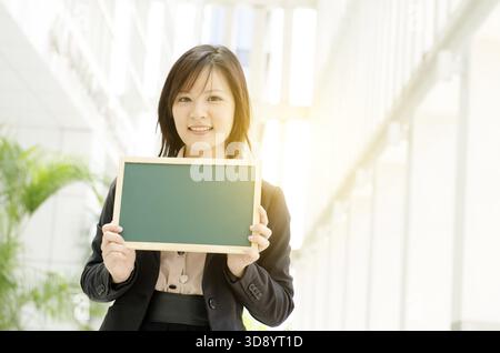 Porträt einer jungen asiatischen Geschäftsfrau, die lächelt und eine leere grüne Tafel hält, in einer Büroumgebung steht, wunderschönes goldenes Sonnenlicht Stockfoto