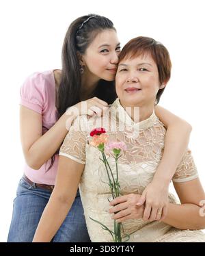 Ältere Mutter mit Nelkenblume, Erwachsene Tochter umarmt und küsst Mutter, isoliert auf weißem Hintergrund. Gemischtes asiatisches Familienporträt Stockfoto