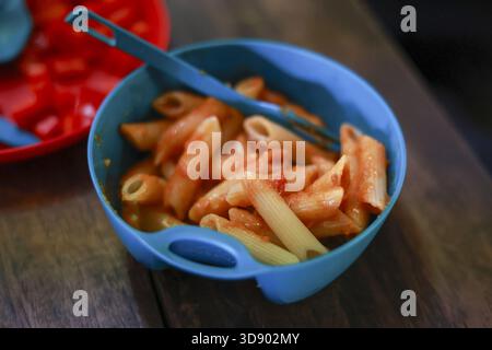 Einfaches hausgemachtes Abendessen mit Penne-Pasta mit Tomatensauce in blauer Plastikschale. Diese angenehme und gesunde Kindermahlzeit steht auf Holztisch, bereit für das Mittagessen Stockfoto
