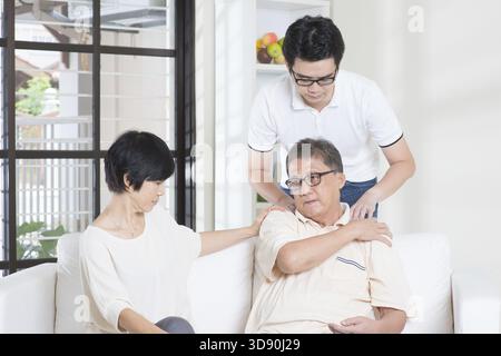 Schulterschmerzen des asiatischen alten Mannes, mit seiner Frau auf dem Sofa sitzend, der Sohn massiert die Schulter des Vaters. Chinesische Familie, Seniorenpensionär im Haus, Lebensstil zu Hause Stockfoto