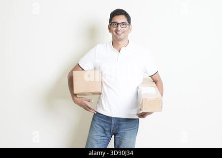 Servicekonzept für Kurierzustellung. Glücklicher Indianer erhielt braune Kästen, die auf einem schlichten Hintergrund standen. Asiatisches, gutaussehendes Männermodell Stockfoto