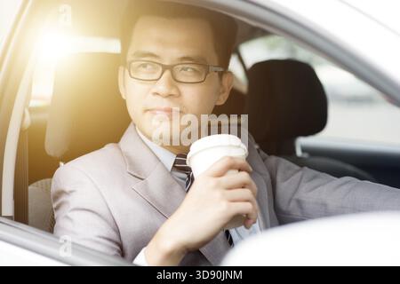 Asiatischer Geschäftsmann trinkt Kaffee, während er mit dem Auto fährt, an einem Morgen mit wunderschönem Sonnenlicht Stockfoto