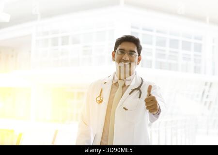 Porträt eines lächelnden asiatischen indischen männlichen Arztes in Uniform Daumen oben, stehend außerhalb des Krankenhausgebäudes, schönes goldenes Sonnenlicht am bac Stockfoto