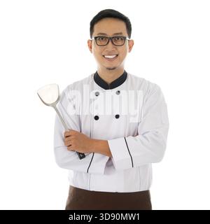 Porträt des asiatischen Küchenchefs, der Spachtel hält, lächelt und isoliert auf weißem Hintergrund steht Stockfoto