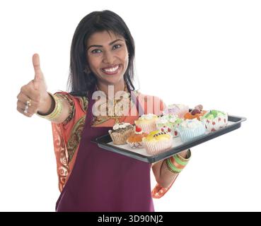 Glückliche, traditionelle Indianerin in Sari, die Brot und Cupcakes backt und den Daumen nach oben zeigt, mit Schürze, die Tablett isoliert auf weiß hält Stockfoto