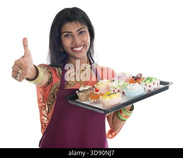 Glückliche, traditionelle Indianerin in Sari, die Brot und Cupcakes backt und den Daumen nach oben zeigt, mit Schürze, die Tablett isoliert auf weiß hält Stockfoto