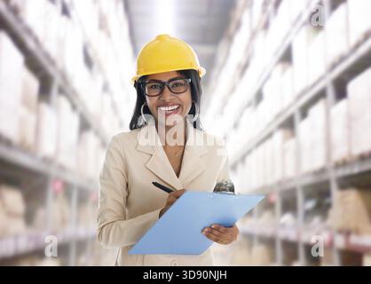 Lächelnde indische Frau, die im Lager mit einem Sicherheitshelm tickt, lächelt glücklich, einen Bericht zu schreiben. Porträt eines schönen asiatischen weiblichen Models, das steht Stockfoto