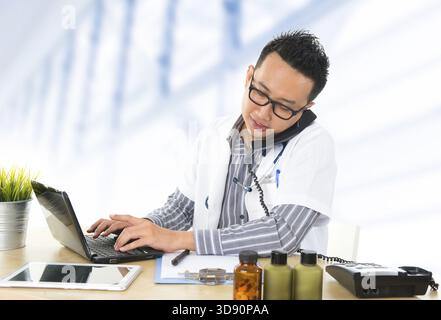 Beschäftigte südostasiatische Ärztin am Telefon und Tippen auf Notizbuch, sitzend an der Rezeption Arbeitstisch im Krankenhaus Stockfoto
