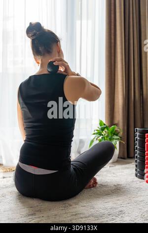 Junge Frau, die zu Hause auf dem Boden sitzt und einen Massageball benutzt, um Nackenschmerzen zu lindern. Selbstpflegeroutine für Muskelverspannungen und Halswirbelsäulengesundheit. Stockfoto