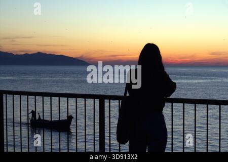 Einsame Silhouette bei Sonnenuntergang über dem Meer Die Silhouette Einer Frau steht auf einem Balkongeländer und blickt auf das ruhige Meer, während die Sonne untergeht Stockfoto