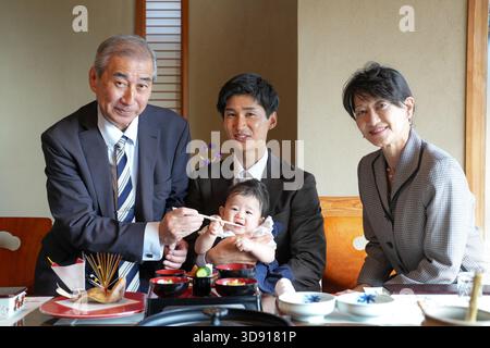 Eine vierköpfige Familie sitzt an einem Tisch mit Essen, einschließlich einer Schüssel Nudeln. Der Mann in der Mitte hält einen Löffel und füttert ein Baby. Szene ist W Stockfoto