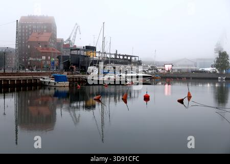 Das Hafenrestaurant und die vertäuten Boote säumen ruhiges Wasser mit roten Bojen und nebeligen Wohnhäusern aus Ziegelsteinen, die im Hintergrund in Helsinki, Finnland, zu sehen sind. Stockfoto