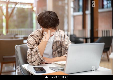 Mann in kariertem Hemd hält oder massiert seinen Tempel vor einem Laptop und Buch, der am Kaffeetisch sitzt. Studieren oder Arbeiten im Freien. Kopfschmerzen oder Migräne Stockfoto