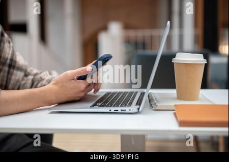 Nahaufnahme eines Mannes, der die Hände hält und das Telefon über Laptop mit Kaffee und Buch ansieht und am Kaffeetisch sitzt. Studieren oder arbeiten, nach draußen gehen. Stockfoto