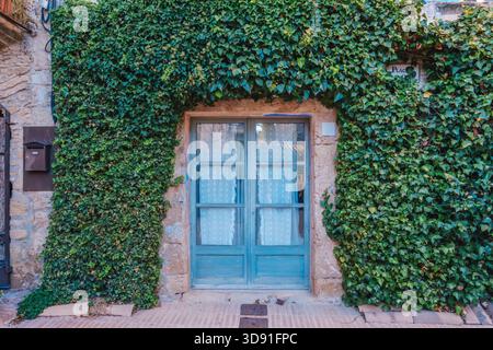 Mittelalterliche Steinfassade mit üppigem grünem Efeu, hellblauer Tür, Bogenfenstern und quadratischem blauem Fensterrahmen in Peratallada Stockfoto