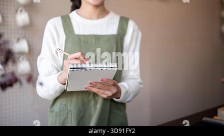 Asiatische Barista-Frau mit grüner Schürze in Notizbuch mit Bleistift, die von einem Gast an der Theke bestellt wird. Café oder Café-Bar, kleine Unternehmen. Stockfoto