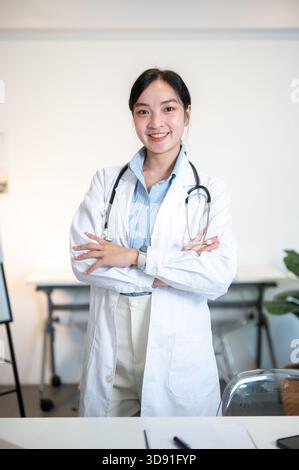 Asiatische Ärztin mit Stethoskop lächelt, während sie mit gekreuzten Armen im Diagnose- oder Untersuchungsraum steht. Krankenhaus oder Klinik, modernes Büro. Stockfoto