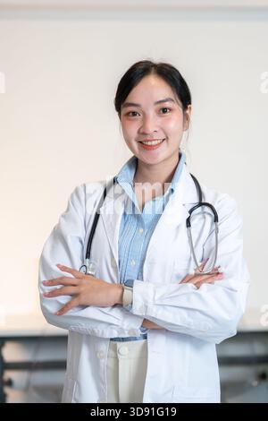 Asiatische Ärztin mit Stethoskop lächelt, während sie mit gekreuzten Armen im Diagnose- oder Beratungsraum steht. Krankenhaus oder Klinik, modernes Büro. Stockfoto