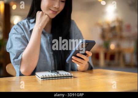 Asiatische Frau, die den Kopf auf der Hand hält und das Telefon ansieht, indem sie eine SMS mit einem Notizbuch auf dem Holztisch des Cafés schickt. Arbeiten oder Studieren. Kühlung Im Freien Stockfoto