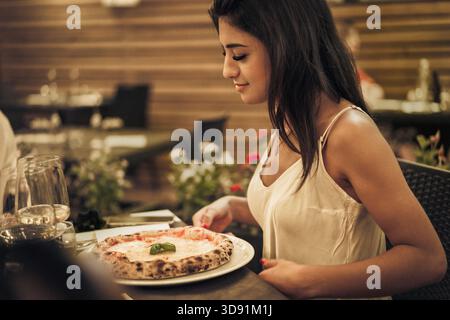 Eine junge Frau sitzt an einem Tisch in einem warmen Restaurant und lächelt, während sie eine leckere Pizza vor sich bewundert. Die Atmosphäre ist einladend, gefüllt mit Stockfoto