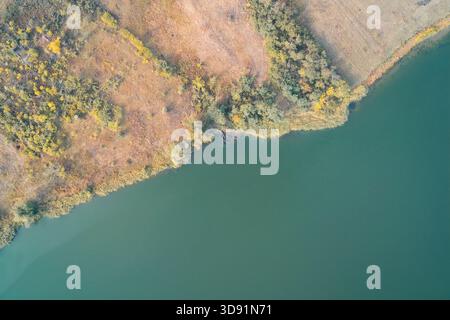 Abstrakter Drohnenschuss, der die diagonale Grenze zwischen einem ruhigen, grünen Wasserkörper und einem strukturierten Flussufer erfasst. Die Mischung aus trockenem Gras, gelben Bäumen und dunklem Wasser bildet einen malerischen natürlichen Hintergrund. Stockfoto