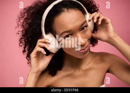 Eine junge Frau mit lockigen Haaren lächelt glücklich, während sie Musik auf ihren Kopfhörern hört. Stockfoto