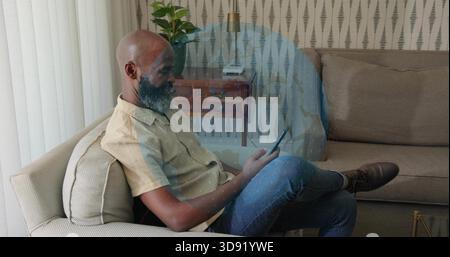 Sitzender Mann mit Tablette auf beigem Sofa zu Hause, mit leichtem Kurzarmhemd und blauer Jeans Stockfoto