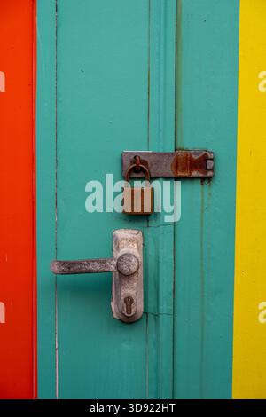 Altes, rostiges Türschloss und Türgriff auf einer hell bemalten Strandhütte. Korrodiertes altes Schloss und Vorhängeschloss mit Haspe und Klammer. Stockfoto
