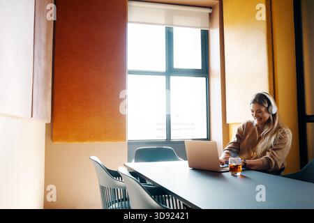 Eine junge Frau sitzt in einem hell beleuchteten Büro und benutzt einen Laptop, während sie Kopfhörer trägt. Das Ambiente ist professionell und einladend, mit Schwerpunkt auf Conce Stockfoto