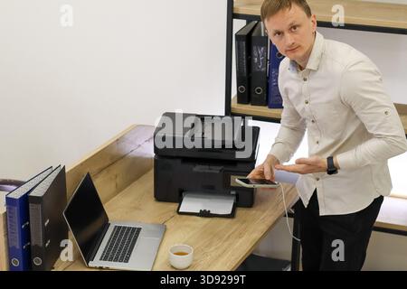 Büromitarbeiter drückt Verwirrung aus, indem sie mit dem Smartphone in der Hand in der Nähe eines schwarzen Druckers auf dem Schreibtisch mit Computer und Ordnern zucken. Stockfoto