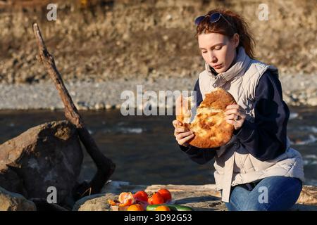 Junge Frau, die auf großen Felsbrocken am Fluss sitzt und traditionelles Fladenbrot mit Blick auf den Bergfluss im Herbst isst Stockfoto