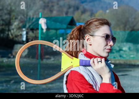 Nahaufnahme einer jungen Frau in sportlicher Kleidung, die einen lebhaften Schläger über der Schulter vor grünem Netz und bergigem Hintergrund hält. Stockfoto