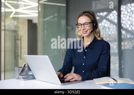 Kundendienstmitarbeiter in einem modernen Callcenter lächelt und tippt auf einem Laptop, während sie ein Headset trägt, und bietet freundlichen, professionellen technischen und verkaufsfördernden Support an ihrem Schreibtisch Stockfoto