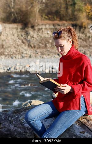 Legere Frau mit rotem Rollkragen sitzt im Kreuz auf Baumstamm, in das Buch mit fließendem Strom und natürlicher Kulisse Stockfoto