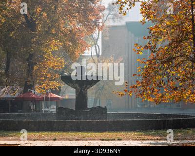 Jerewan Armenien 11.25.2025. Ein Brunnen in einem Park mit einem roten Schirm in der Nähe. Der Brunnen ist von Bäumen umgeben und hat eine Vogelstatue in der Mitte. Stockfoto