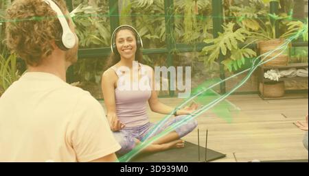 Sitzende Frau mit Fliedertank und Leggings, die auf Matte im Sonnenraum mit Kopfhörern und Weihrauch meditiert Stockfoto