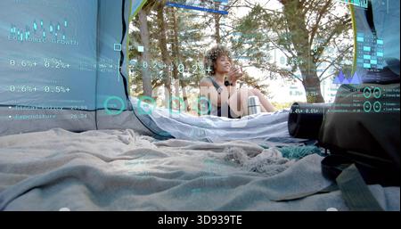 Eine sitzende Frau schlürft einen Becher und überprüft das Telefon im Zelt in Shorts, mit Becher und AR-Überzügen Stockfoto