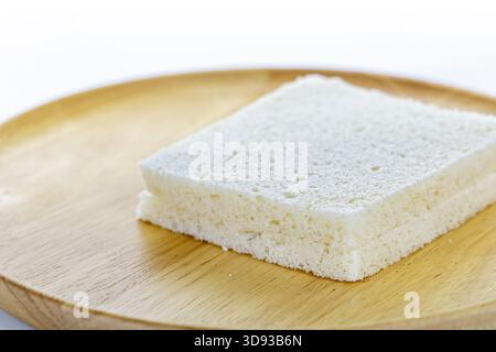 Aus nächster Nähe eine quadratische Scheibe Weißbrot auf einem Holzteller, die die weiche, poröse Textur des Brotes vor einer einfachen, verschwommenen Struktur hervorhebt Stockfoto