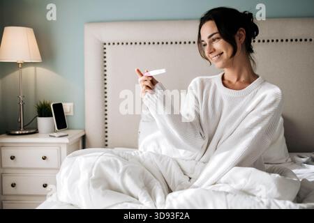 Glückliche junge Frau in weißem Pullover, die auf dem Bett sitzt, mit Aufregung den Schwangerschaftstest hält, umgeben von weicher Bettwäsche und warmem Licht, die eine Freude vermittelt Stockfoto