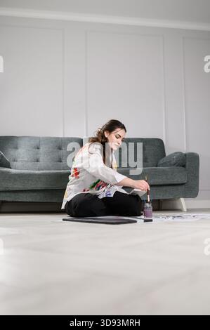 Talentierter Künstler in lackierten Hemdfarben, während er auf dem Boden in der Nähe des Sofas sitzt. Frau bastelt Bodenmalerei in hellen, modernen Räumen. Kreativer sessi Stockfoto